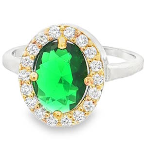 Cette bague élégante allie un design classique à une touche de contraste moderne. La bague est confectionnée en argent 925/000 rhodié, offrant une finition brillante et épurée. L'anneau est fin et lisse, ce qui permet de mettre l'accent sur la tête imposante de la bague. Le centre de la bague présente un jeu de couleurs sophistiqué. Tandis que le corps de la bague est en argent, les griffes retenant la pierre centrale et le liseré intérieur du halo sont dorés, créant un effet bicolore très tendance. Elle arbore une magnifique pierre verte éclatante (évoquant une émeraude) de taille ovale. La pierre est facettée pour capturer la lumière et révéler des reflets profonds et cristallins. La pierre centrale est entourée d'un halo de petits cristaux blancs (oxydes de zirconium). Ces pierres apportent un éclat supplémentaire et renforcent l'aspect luxueux de la pièce, rappelant le style des bagues d'entourage classiques.